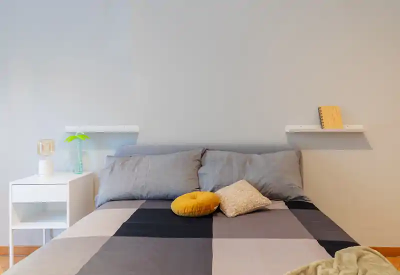 Bright, modern bedroom with a centrally framed double bed, grey pillows and decorative cushions creating a clean, inviting look. Minimal bedside table and shelving emphasize a neat, contemporary style ideal for rental photos.