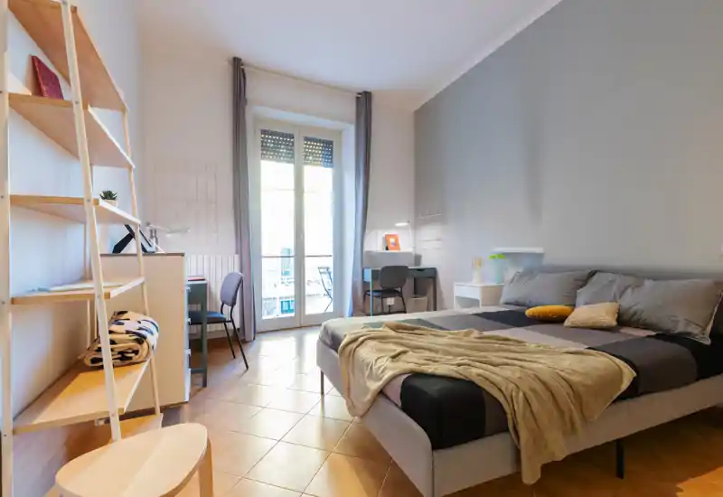 Bright, modern bedroom with a double bed, soft throw and pillows, desk and shelving, plus a balcony door letting in natural light — ideal for comfortable stays.