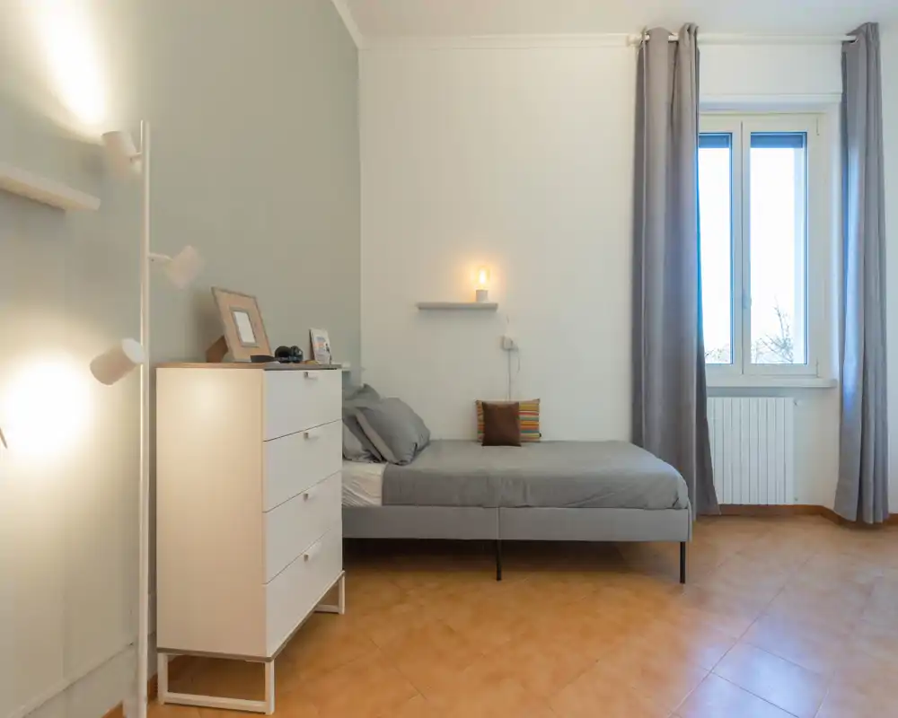 Chambre minimaliste et soignée avec un lit double gris, du linge de lit doux, une étagère de chevet avec une lampe chaude et une fenêtre lumineuse encadrée de rideaux clairs.
