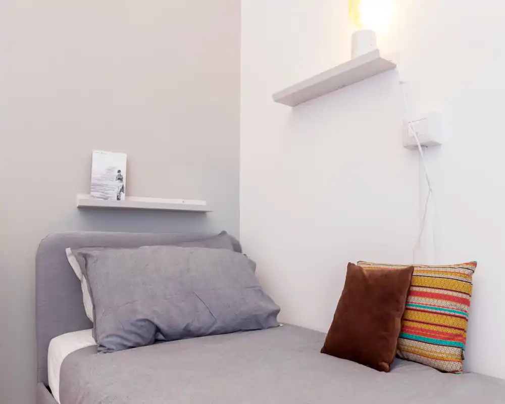Coin chambre simple, soigné et bien décoré avec lit bien fait, linge de lit gris et coussins colorés. Une étagère murale et une lampe apportent une touche moderne et chaleureuse, idéal pour une annonce locative.
