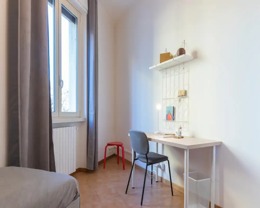 Coin chambre simple et lumineux avec un lit partiellement visible à gauche, un bureau et une chaise près de la fenêtre et des étagères décoratives minimalistes.