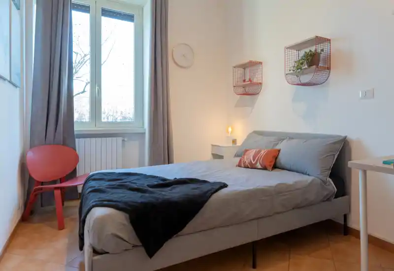 Bright, modern bedroom with a comfortable double bed, soft linens and a decorative cushion. Natural light from the window and simple, tasteful shelving create a calm, welcoming atmosphere.