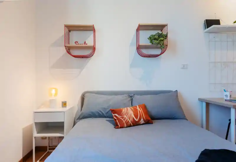 Bright, modern bedroom with a neatly made double bed dressed in grey linens, a decorative orange cushion, bedside table with lamp and minimalist wall shelving — ideal for short-term rental photos.
