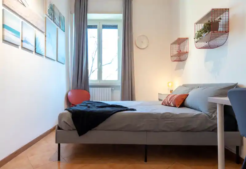 Bright, well-composed double bedroom with neutral linens, decorative cushions and a bedside lamp; large window provides natural light and a calm, modern atmosphere.