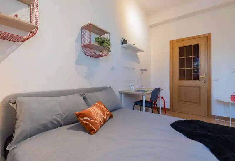 Bright, well-styled bedroom with a large grey bed, decorative cushion and a small desk by the door — comfortable and ready for a guest.