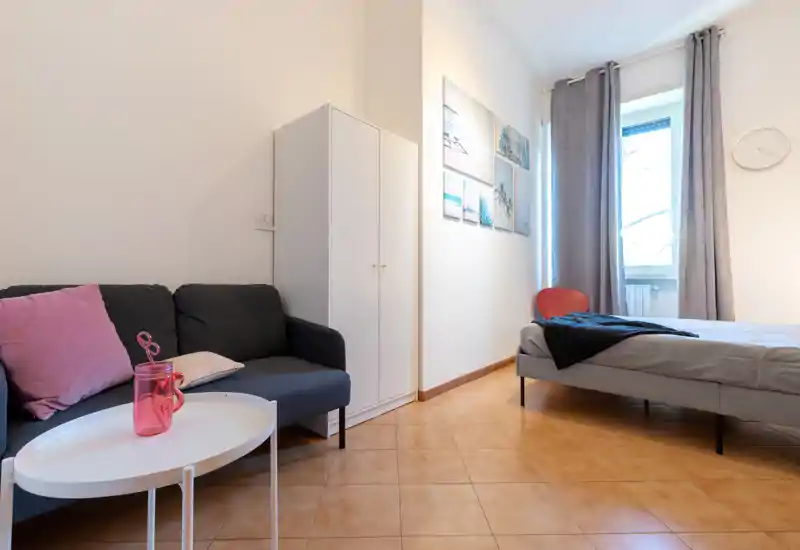 Bright, compact bedroom with a double bed and a small seating area. Neutral décor, natural light from a large window and tasteful wall art make the space feel comfortable.