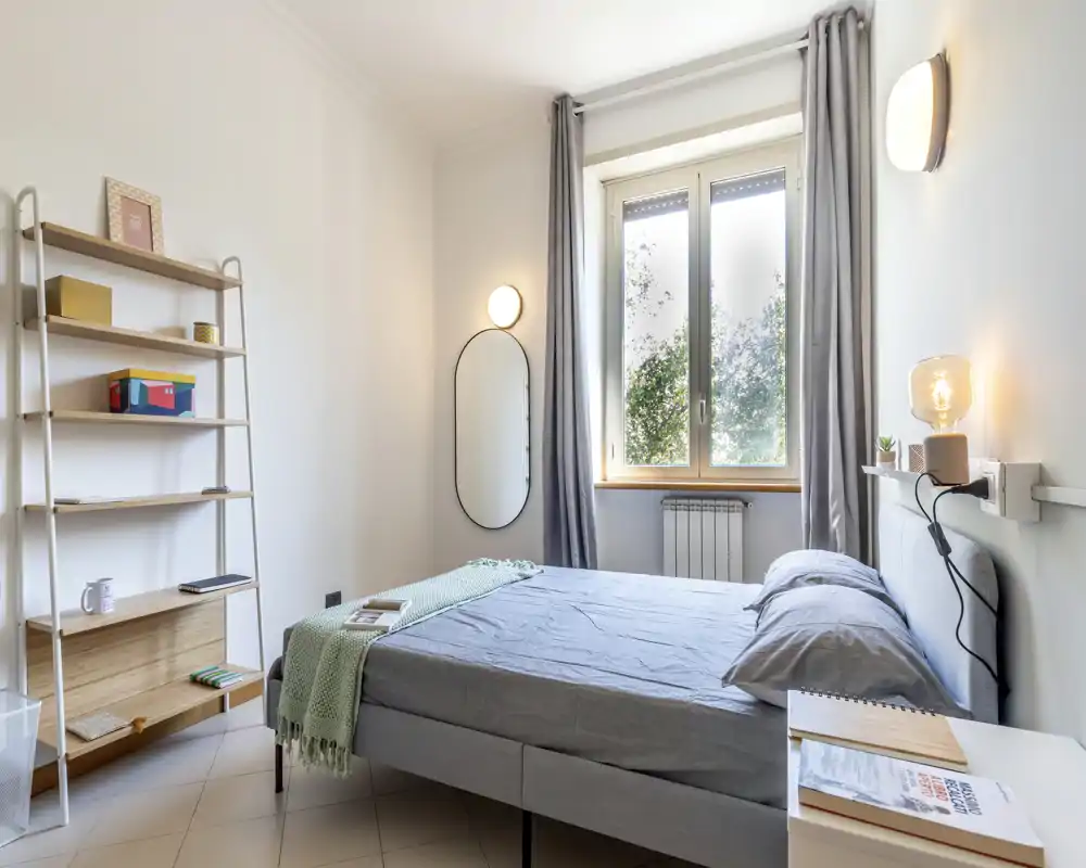Dormitorio luminoso y moderno con cama doble bien hecha, ropa de cama gris, luces de mesita y una gran ventana que aporta luz natural.