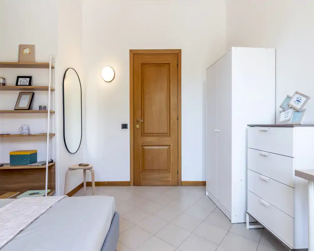 Bright, neatly styled bedroom corner with a visible bed edge, white storage furniture and warm wooden accents creating a calm, modern atmosphere.
