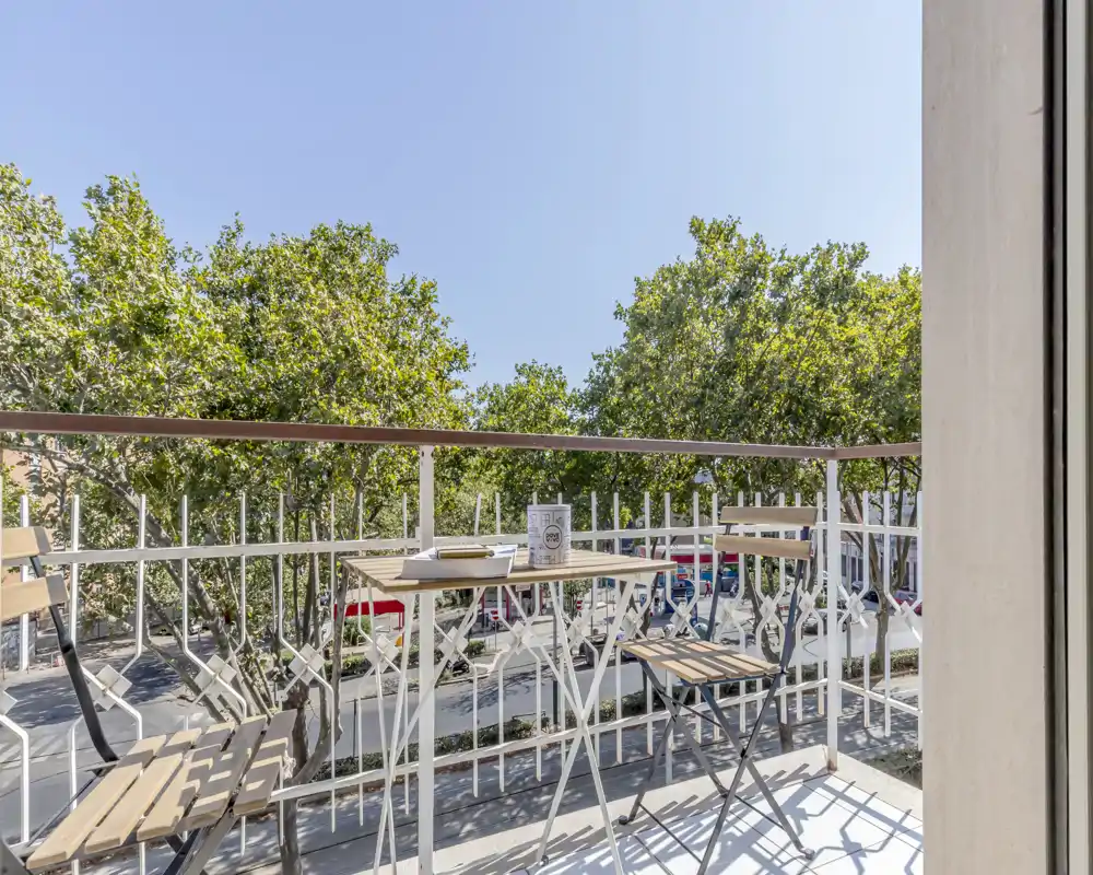 Sunny balcony with a small bistro table and two folding chairs, overlooking treetops and a quiet street — bright, airy spot for morning coffee.