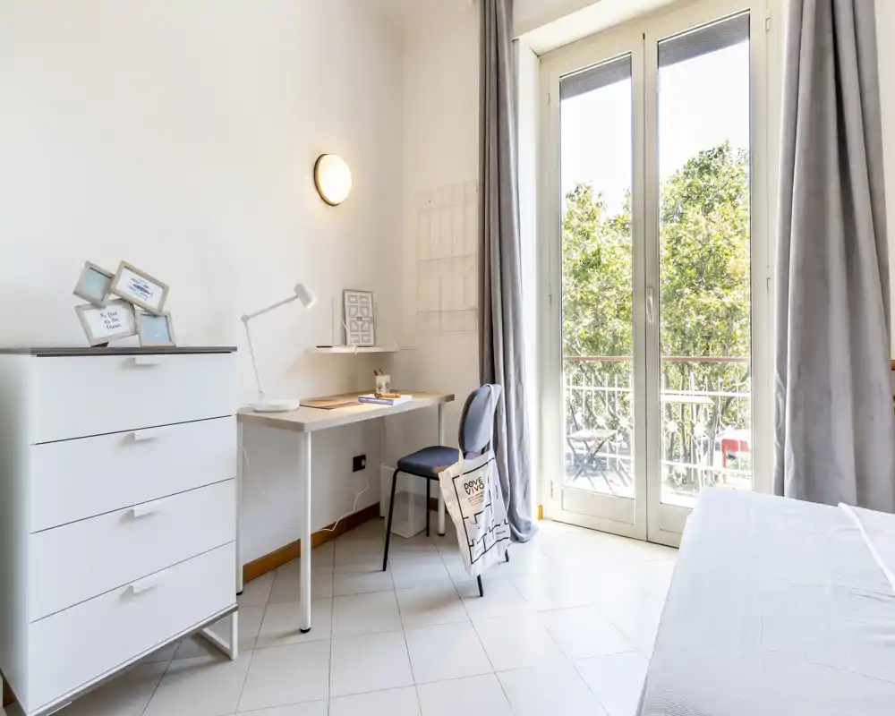 Bright, tidy bedroom with a partially visible bed, a desk and chest of drawers next to a large balcony door that floods the room with natural light.