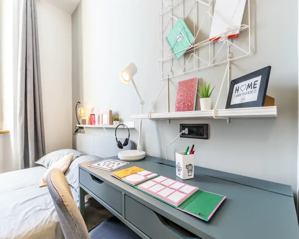 Dormitorio moderno y luminoso con la cama bien cuidada y una zona de escritorio funcional, ideal para estudio con espacio para trabajar y descansar. Líneas limpias, textiles suaves y estanterías ordenadas transmiten calma y confort.