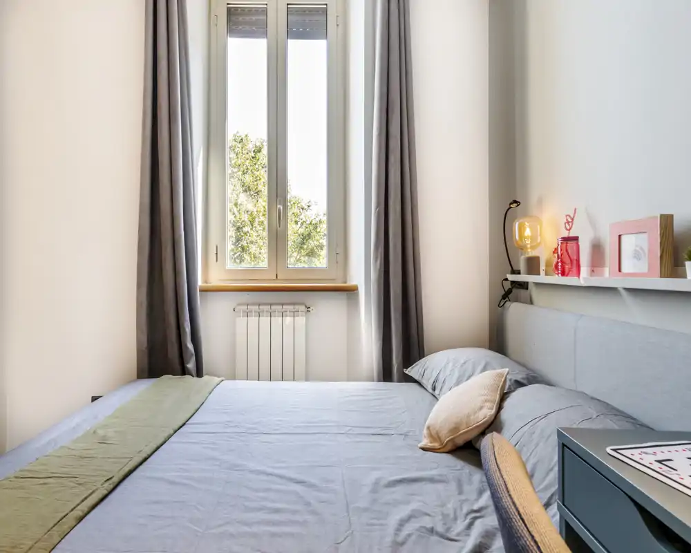 Dormitorio moderno y luminoso con cama doble bien hecha bajo una ventana grande; textiles suaves, lámpara de noche y decoración en la balda crean una atmósfera acogedora.
