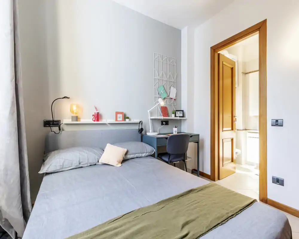 Dormitorio luminoso y bien compuesto con cama de tamaño completo, lámpara de noche y una zona de escritorio junto a la puerta que conduce al baño en suite.