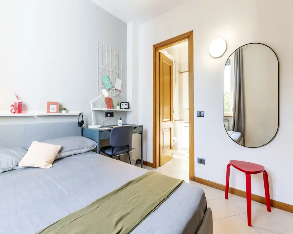 Dormitorio moderno y luminoso con una cama amplia, una repisa de mesilla y un escritorio compacto. Ropa de cama de tonos neutros, espejo decorativo y luz natural crean un ambiente fresco y acogedor ideal para estancias cortas.