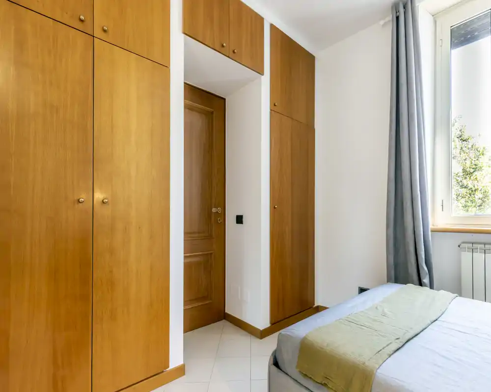 Dormitorio luminoso con armarios empotrados de madera, cama doble parcialmente visible con ropa de cama neutra y una gran ventana que aporta luz natural.