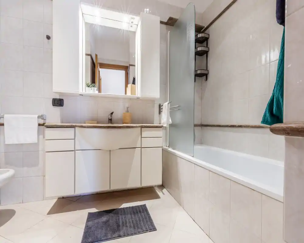 Baño moderno y luminoso con bañera y lavabo integrado; espejo mural y mampara de vidrio, azulejos claros y ambiente ordenado.