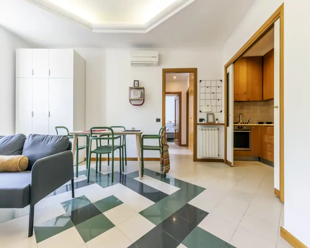 Zona de estar moderna y luminosa con mesa de comedor compacta, sillas verdes y un sofá cómodo; vistas abiertas a la cocina y al dormitorio para un diseño práctico y aireado.