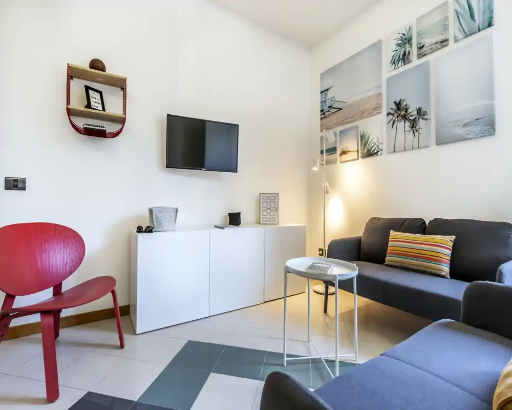 Sala de estar moderna y luminosa con sofá oscuro, cojín colorido, mesita redonda y televisor de pared; cuadros con temática de playa y una silla roja completan el ambiente acogedor.