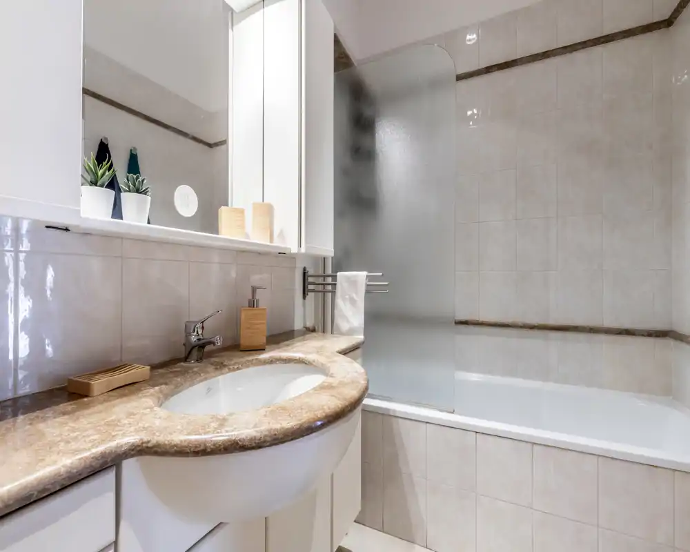 Baño luminoso y limpio con encimera efecto mármol, lavabo integrado y bañera con mampara de vidrio esmerilado; instalaciones modernas y azulejos neutros que crean un ambiente fresco.