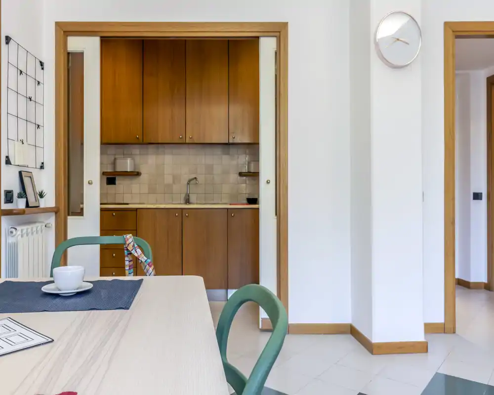 Área de comedor luminosa y ordenada que se abre a una cocina compacta con armarios de madera cálida; una mesa clara con individual y sillas crea un espacio agradable para comer.