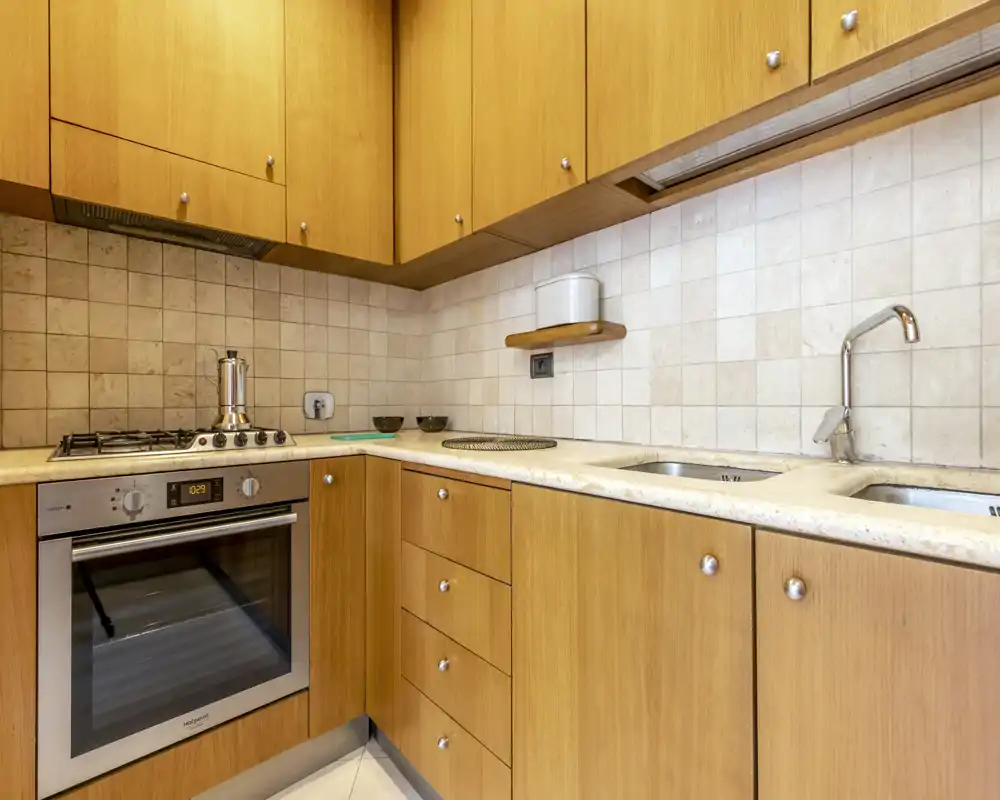 Cocina compacta en madera cálida con horno integrado, placa de gas y fregadero doble. Salpicadero de azulejos neutros e iluminación bajo los armarios que crean un espacio de cocina práctico y ordenado.