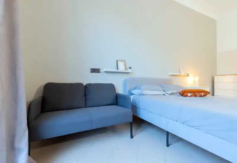 Camera da letto moderna e luminosa con un comodo letto matrimoniale in lenzuola chiare e un piccolo divano accanto; decor minimal e una lampada da comodino calda creano un’atmosfera rilassante.