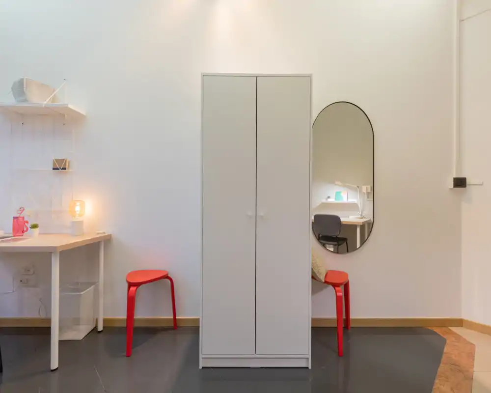 Neat, minimal living area with a central wardrobe, two red stools, a small desk with lamp and wall mirror — bright and simply styled for a compact space.