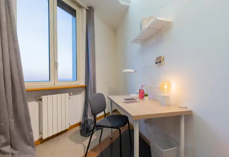 Compact workspace by a window with a simple desk, chair, shelving and a warm desk lamp. Natural light and soft curtains create a calm, functional corner ideal for work or study.