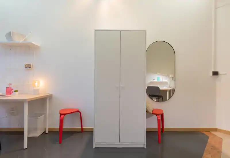 Neat, minimal living area with a central wardrobe, two red stools, a small desk with lamp and wall mirror — bright and simply styled for a compact space.