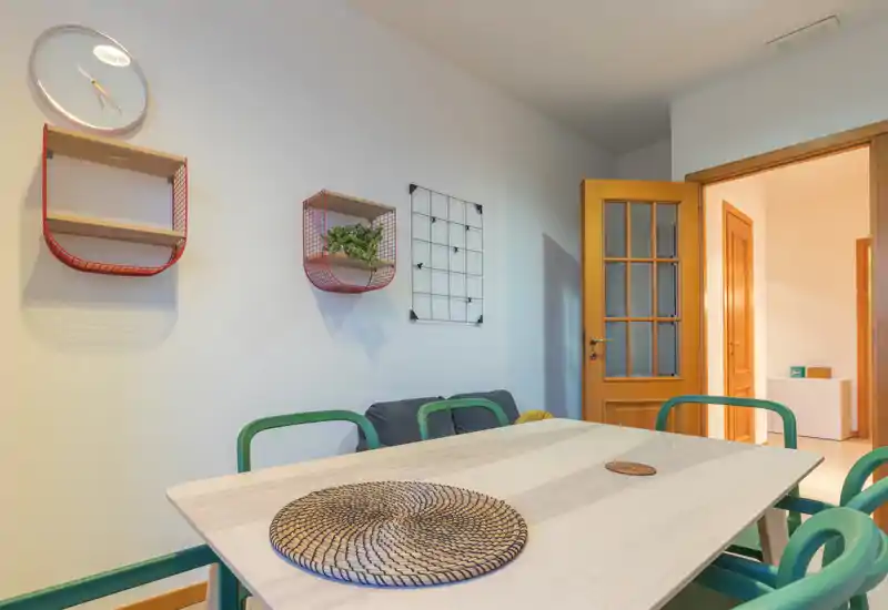Bright, styled dining area with a light wood table, green chairs and decorative wall shelving — a welcoming spot for meals or remote work.