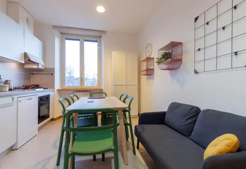 Bright, open-plan living area with a compact kitchen and dining table. The space features a comfortable dark sofa, green dining chairs and modern kitchen appliances — ideal for a small apartment.