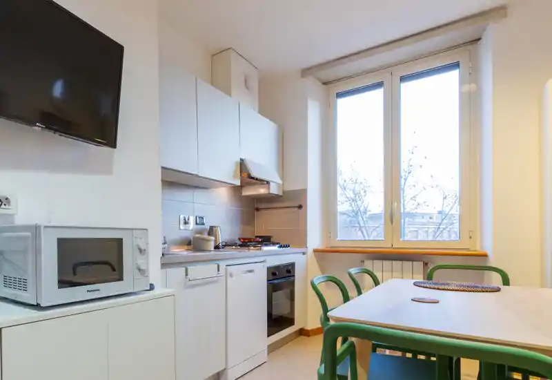 Bright, modern kitchenette with built-in oven, stovetop and dishwasher. A dining table with green chairs sits by a large window, creating a welcoming space for meals.