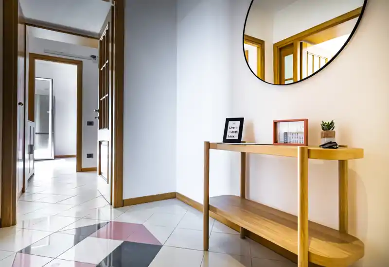Entrée moderne et lumineuse avec une console en bois, un miroir rond et un cadre décoratif ; sol carrelé à motif géométrique et accès clair aux pièces adjacentes.