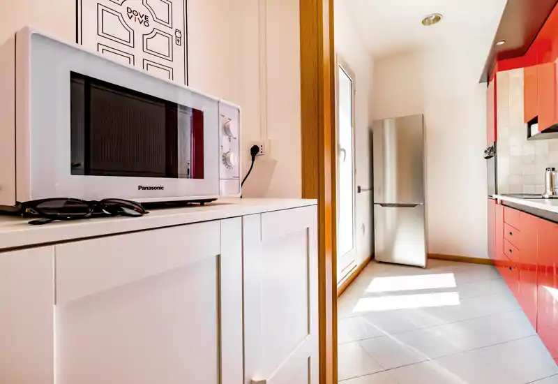 Cuisine moderne et lumineuse avec placards rouges, réfrigérateur inox et micro‑ondes sur un buffet blanc ; carrelage propre et lumière naturelle depuis la porte créent un espace agréable.
