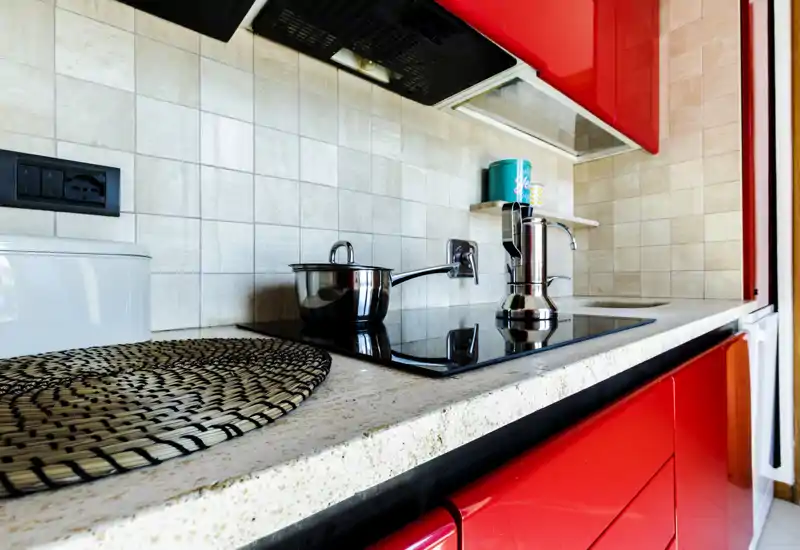 Kitchenette moderne et lumineuse avec plaque électrique design, casserole en acier inoxydable et moka sur un plan de travail clair encadré par des placards rouges laqués.