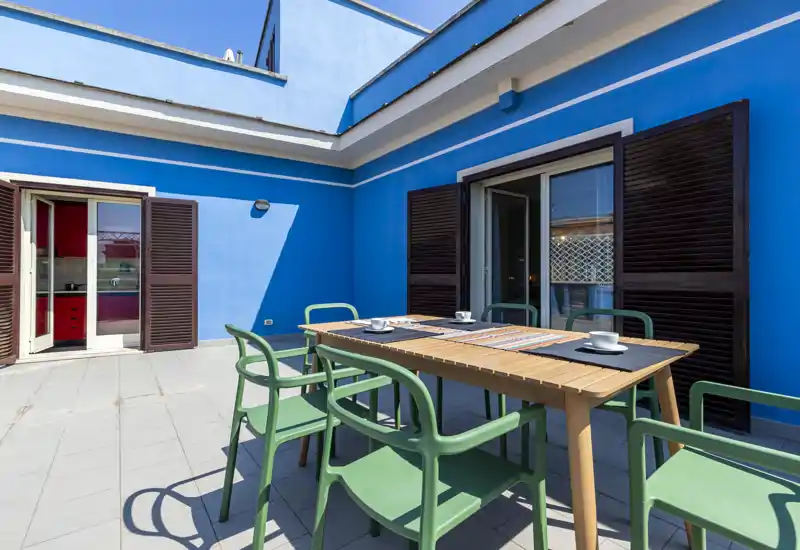 Terrasse privée ensoleillée avec une table en bois et des chaises vertes modernes, idéale pour les repas en extérieur. Les murs bleu vif et les volets offrent une ambiance chaleureuse et pétillante.