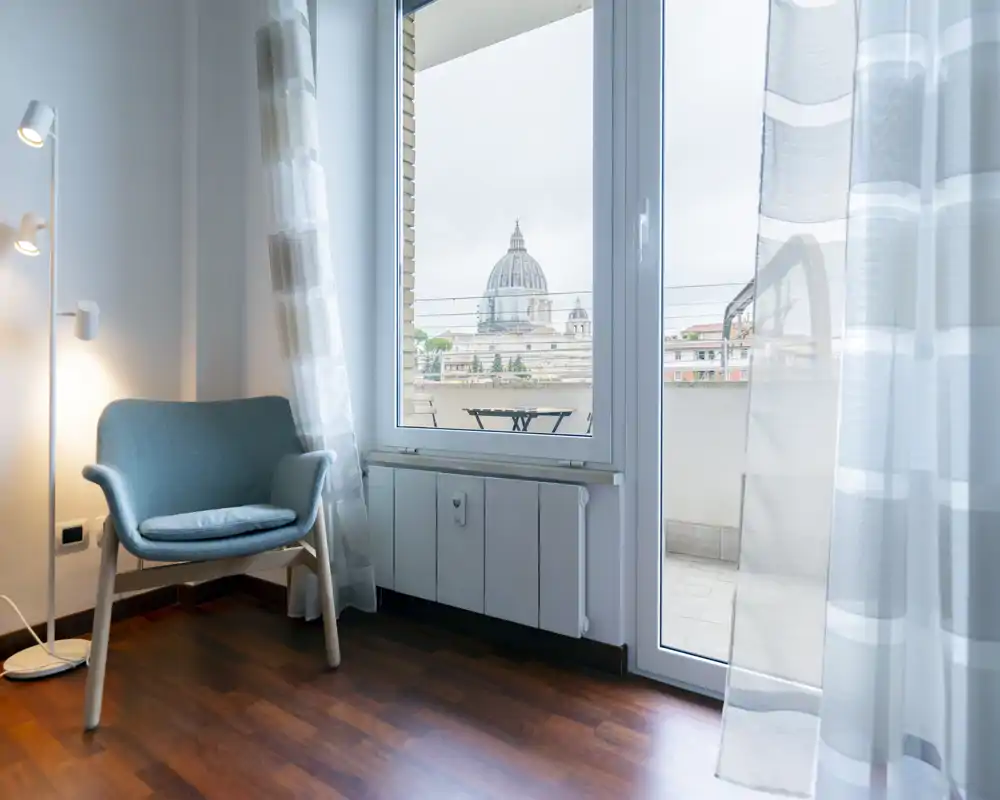 Small bright living corner with a comfortable armchair, standing lamp and large window/door opening to a balcony with city views. Neutral tones and sheer curtains create an airy, modern feel ideal for relaxing or reading.