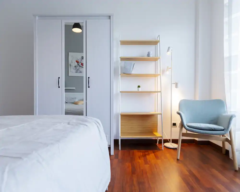Bright, modern bedroom with a white bed in the foreground, a mirrored wardrobe, open shelving and a cozy armchair by the window. Clean minimalist styling and warm wooden floors create a calm, inviting atmosphere.