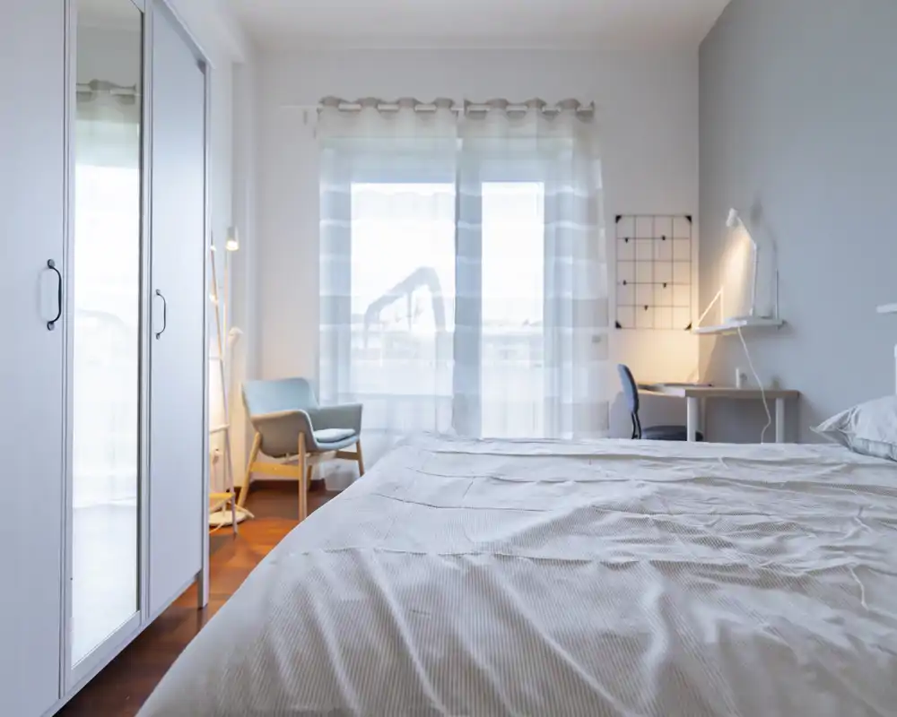 Bright, modern bedroom with a large bed in the foreground, sheer curtains letting in soft natural light, a cozy armchair and a small desk — ideal for restful stays or remote work.