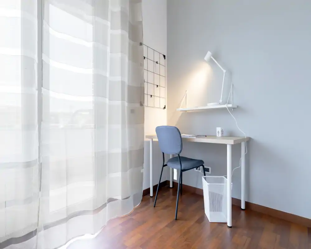 Bright, minimal corner workspace in a living area with a desk, chair, desk lamp and sheer curtains letting in natural light.