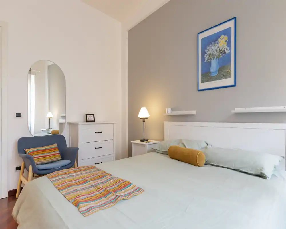 Camera da letto luminosa e ben arredata con un ampio letto matrimoniale, lampada da comodino, cuscini accoglienti e una poltrona vicino a uno specchio alto.