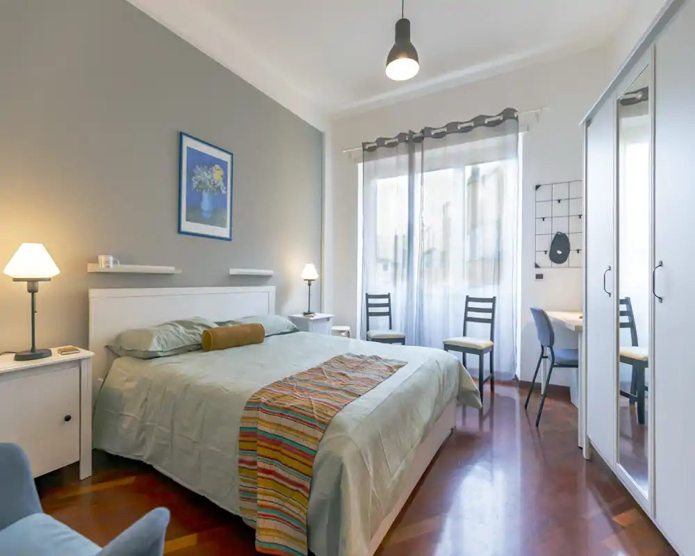 Camera matrimoniale luminosa e curata, con comodo letto, comodini e illuminazione soffusa. Ampia finestra con tende leggere che illumina l’ambiente; armadio con ante specchiate e una scrivania per maggiore praticità.