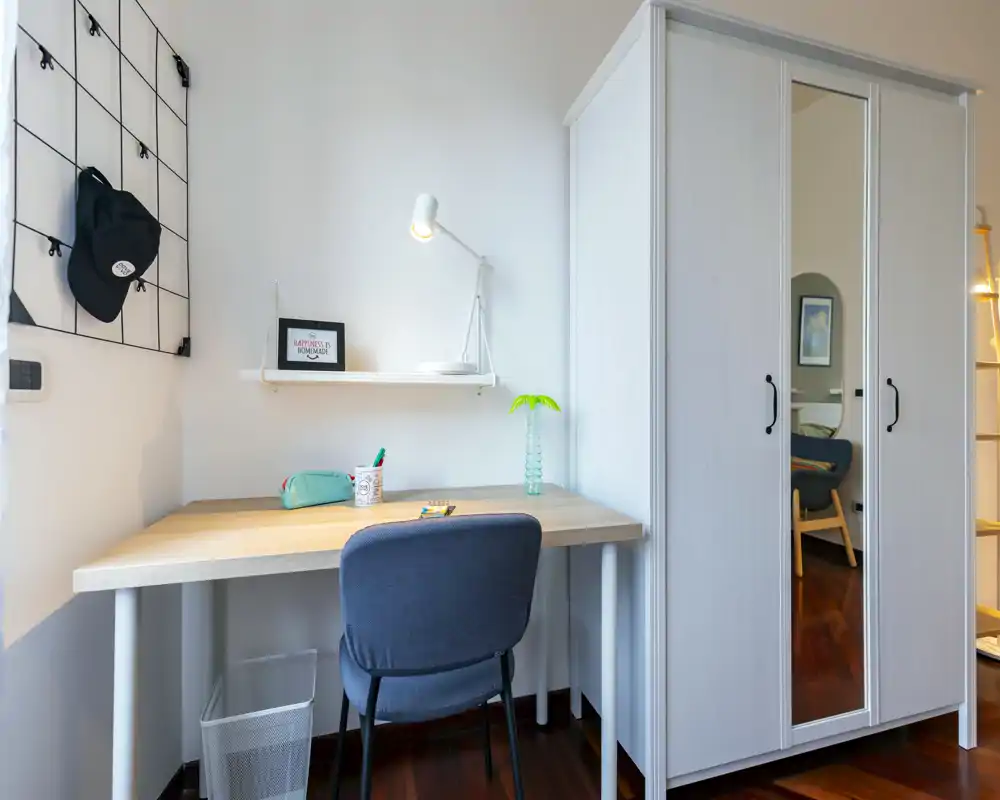 Angolo camera compatto con scrivania ordinata, sedia imbottita e armadio con specchio. Arredo pulito in stile nordico, luminoso e funzionale per lavoro e riposo.