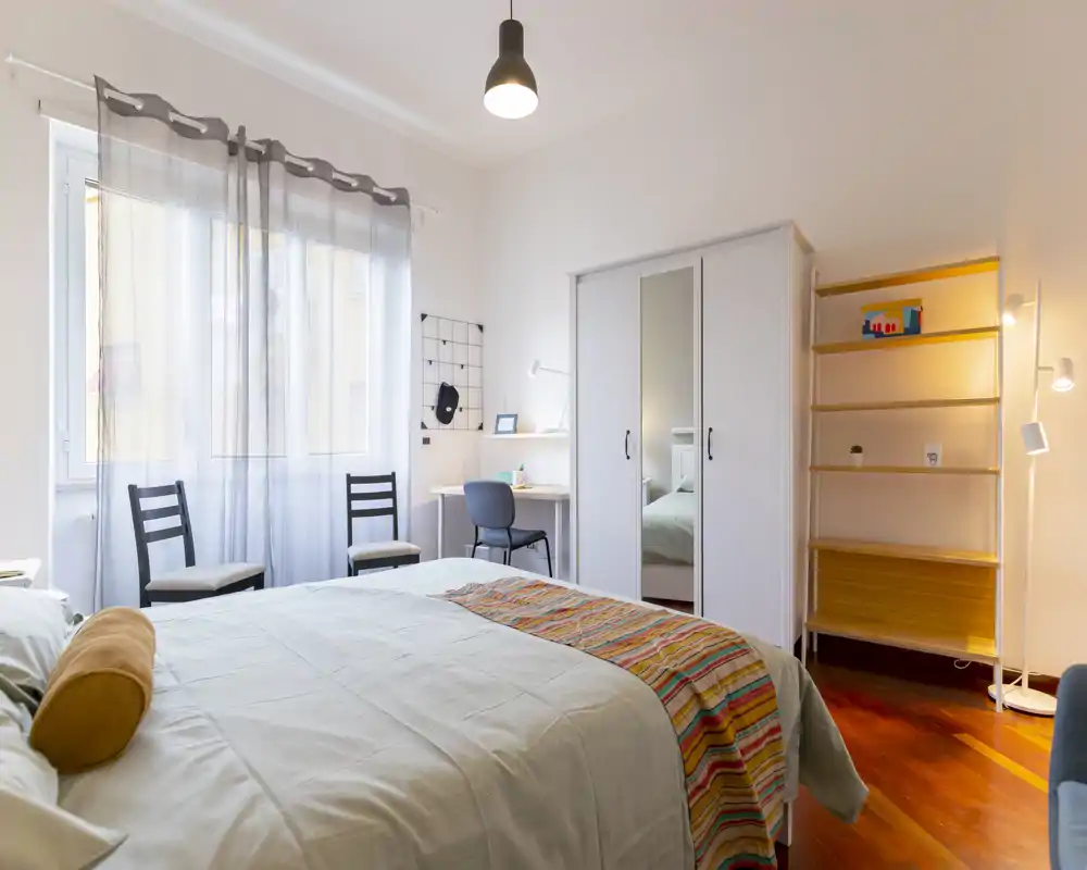 Camera da letto luminosa e ben arredata con letto matrimoniale in primo piano, armadio con specchio, scrivania vicino alla finestra e pavimento in legno caldo.