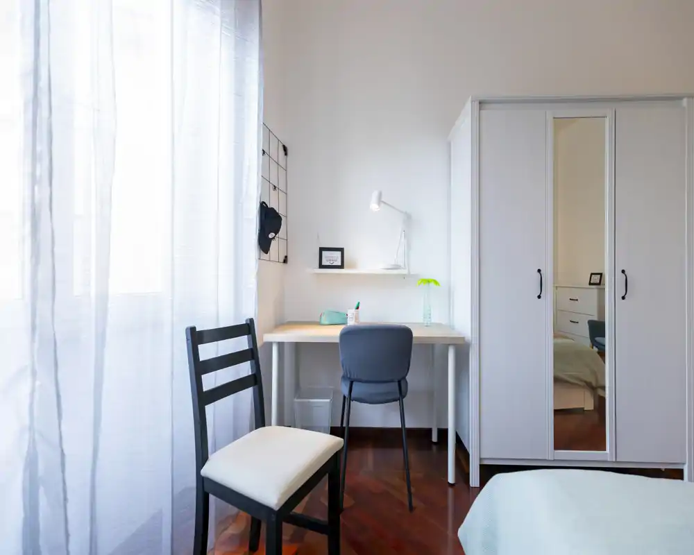 Angolo camera luminoso e minimal con scrivania, armadio con porta a specchio e letto parzialmente visibile. Perfetto per annunci che evidenziano una postazione di lavoro tranquilla e spazio per riporre.