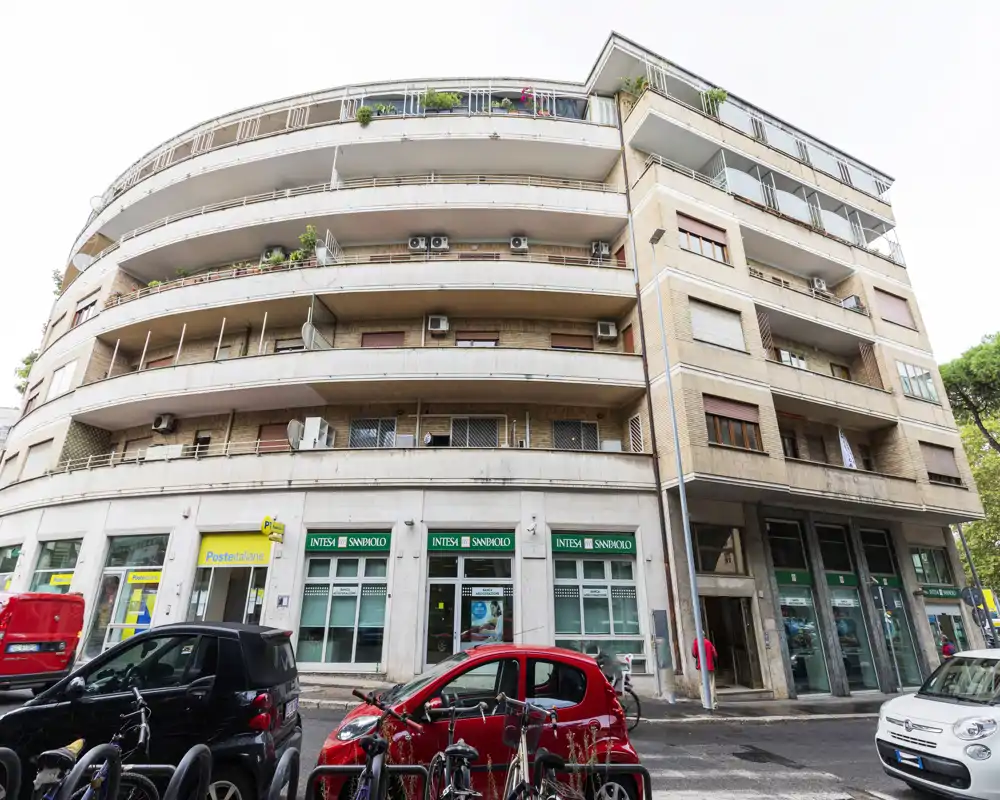 Street-level exterior of a multi-story mixed-use building with balconies, retail units on the ground floor and parked cars in front. The façade and ground-floor shops are clearly visible — useful for showing the building’s frontage and neighborhood context.