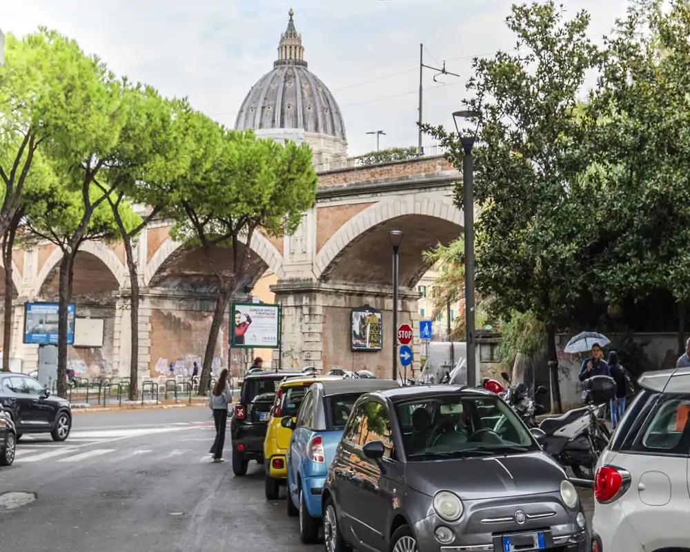Street view with parked cars and an arched viaduct in front of a domed landmark; bright daylight and leafy trees frame the urban scene.