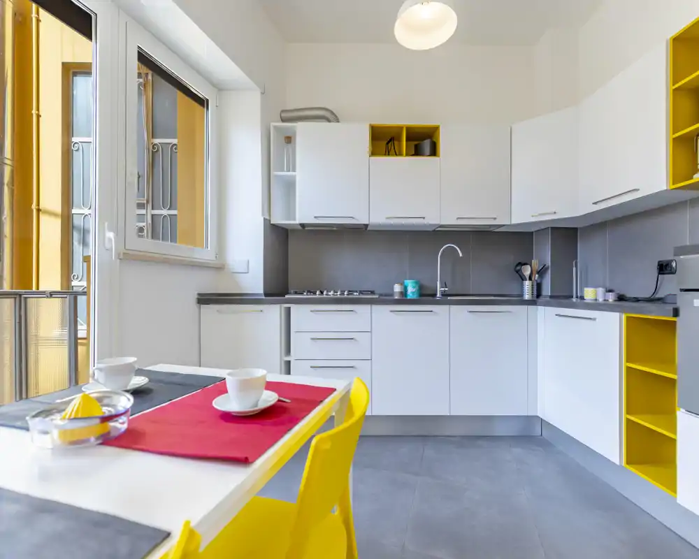 Modern bright kitchen with white cabinets, integrated countertops and yellow accents; small dining table set by a window provides a cozy breakfast corner.