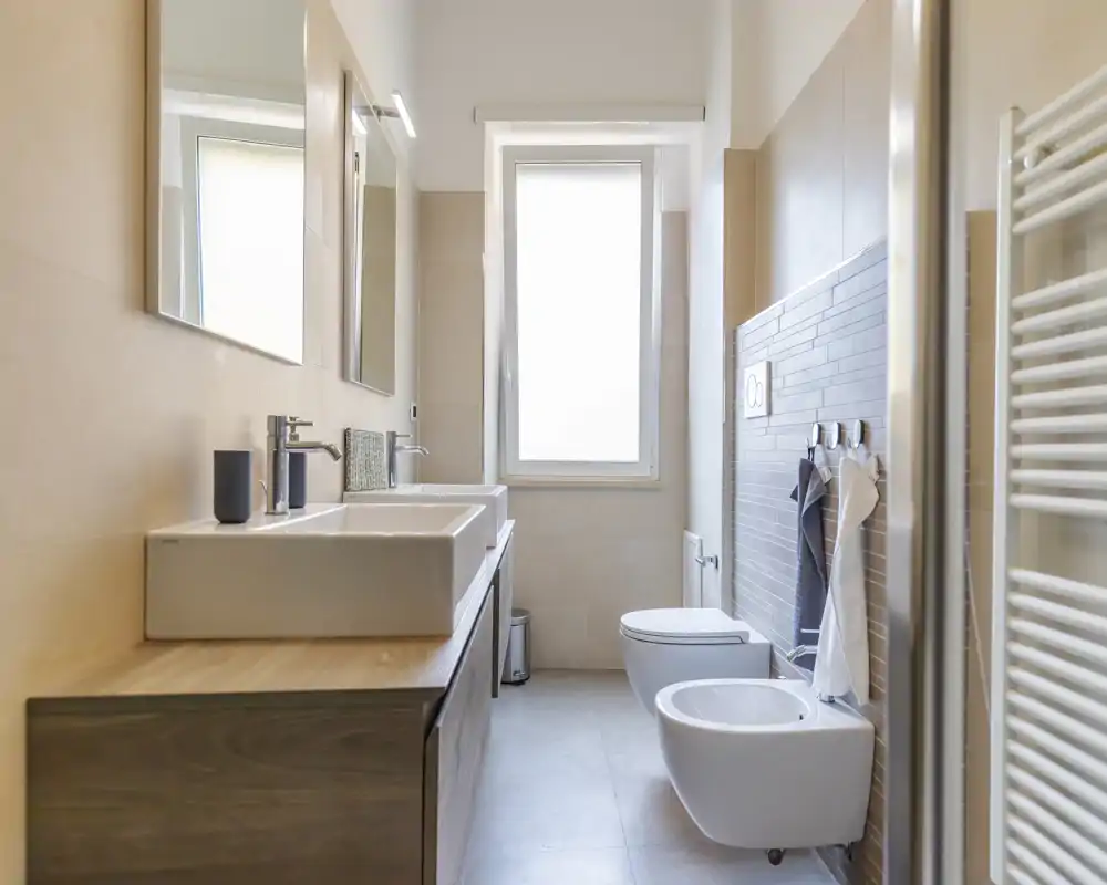 Modern, bright bathroom with twin rectangular sinks, wall-mounted bidet and toilet, large mirror and a window providing soft natural light.