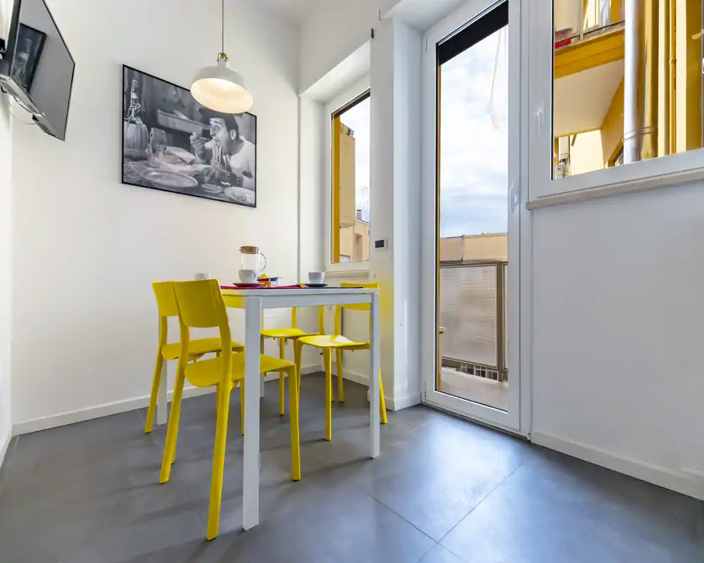 Small modern dining corner with a white table and four bright yellow chairs next to a glass door letting in natural light; simple tableware and a framed black-and-white print add a stylish, inviting touch.